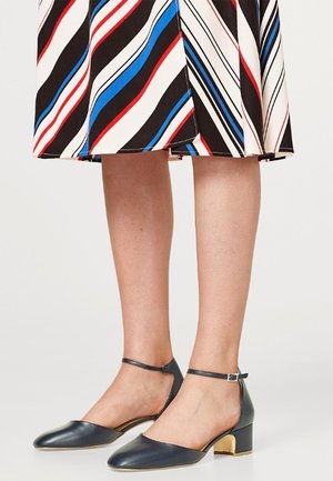 Jambes portant une jupe rayée et des chaussures à talons carrés bas bleu marine avec des brides autour de la cheville sur un fond clair.