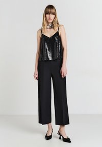 Donna che indossa un top in paillettes nere, pantaloni neri a gamba larga, décolleté neri con slingback e un grande choker a forma di fiore metallico, in piedi contro uno sfondo semplice.