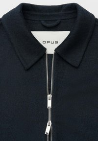 Giacca nera con cerniera frontale, realizzata in tessuto strutturato, con colletto a punta e doppia cerniera con tiretti metallici. Etichetta del marchio visibile all'interno.