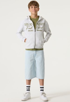 Grijze hoodie met rits en groene tekst, olijfgroen shirt eronder, lichtblauwe knielange korte broek, witte sneakers en zwart-witte gestreepte sokken.