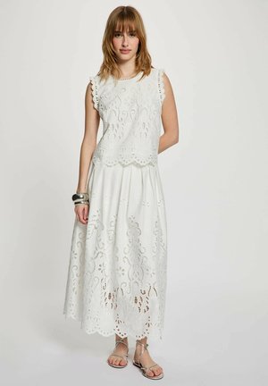 Kvinde stående iført en hvid ærmeløs øjet blonde top og matchende langt skørt med sølv sandaler og to sølv armbånd.