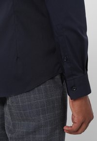 Camisa de algodón azul marino con mangas largas, que presenta puños con botones, combinada con pantalones de cuadros grises. Textura suave, diseño nítido.