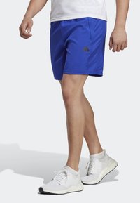 Person som bär blå sportiga shorts med Adidas-logotyp och vita löparskor stående på en vit bakgrund.