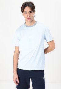 Tricou din bumbac de culoare albastru deschis, cu guler rotund și guler în dungi. Prezintă un logo circular texturat în centrul său. Mânici scurte.