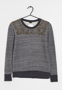 Suéter gris con una tela texturizada, con un escote redondo y tachuelas decorativas en los hombros. Mangas largas y puños acanalados.