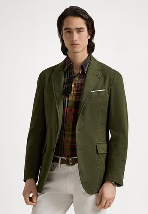 Green blazer with a textured finish, two front pockets, worn over a multicolored plaid shirt. White pocket square accent, minimalistic design.