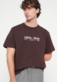 Σκούρο καφέ βαμβακερό t-shirt με κοντά μανίκια, με λευκή εκτύπωση κειμένου "calvin klein new york city" στο στήθος. Casual εφαρμογή.