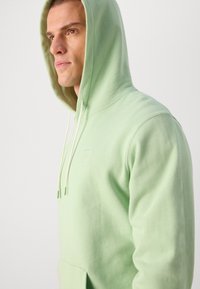 Hoodie vert menthe avec cordon de serrage, présentant une poche avant et un logo discret sur la poitrine. Tissu doux et coupe décontractée.