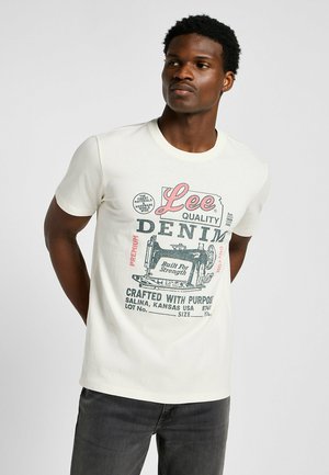 Una camiseta de algodón blanca con un diseño gráfico retro que presenta "Lee Quality Denim" y una ilustración de una máquina de coser en impresión roja y negra.