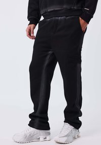Hombre con pantalones joggers negros con logo lateral y zapatillas Nike blancas, de pie con una mano en el bolsillo frente a un fondo blanco.