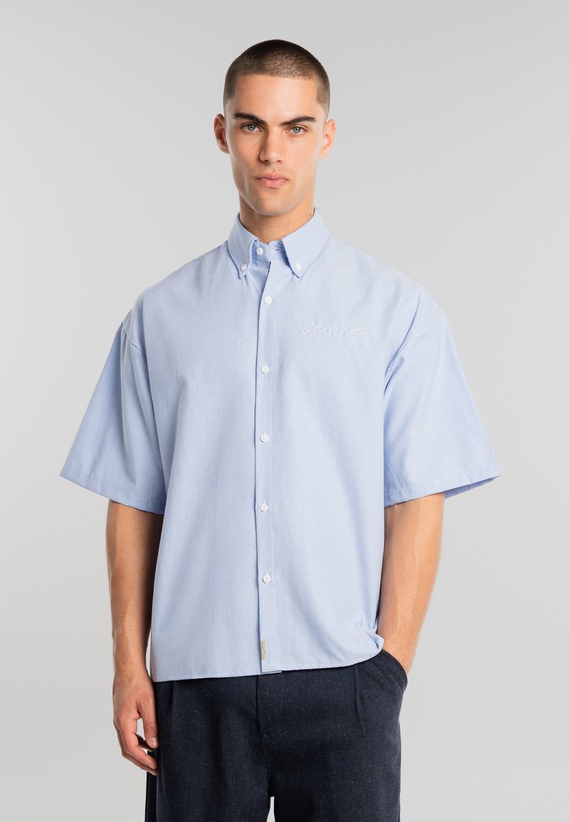 Ung mand iført en lyseblå kortærmet skjorte med knapper og krave, stående foran en ensfarvet lysegrå baggrund.