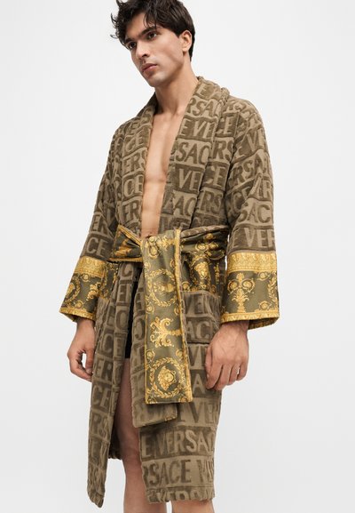 Άνδρας που φοράει ένα καφέ ρόμπα Versace με χρυσή ζώνη και μανσέτες με σχέδιο, στέκεται μπροστά σε έναν απλό λευκό φόντο.