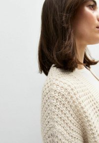 Beige stickad cardigan med ett texturerat mönster, öppen frontdesign och ribbade kanter, som mjukt hänger över axeln.