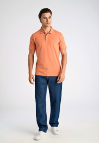 Polo a maniche corte in pesca, con colletto e patta bottoni, abbinato a jeans blu e sneaker bianche su uno sfondo chiaro.