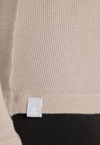 Beige ristunud kangas, mille allosas on sile must osa. Esiplaanil valge silt logoga õmbluse juures, rõhutades tekstuuri ja värvide kontrasti.