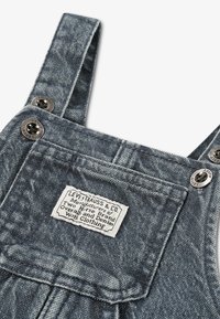 Gros plan sur une salopette en denim bleu avec poche avant et boutons métalliques, avec une étiquette Levi Strauss & Co. cousue sur la poche.
