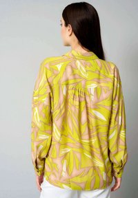 Langärmlige Bluse in leuchtendem Gelb mit abstraktem grünen und pinken Muster. Verfügt über eine Knopfleiste am Rücken und einen weichen, strukturierten Stoff.