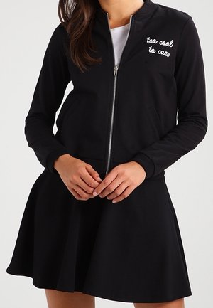 Vrouw die een zwart jack met rits draagt met de tekst "too cool to care" op de borst en een zwarte uitlopende rok, handen voor zich samengevouwen.