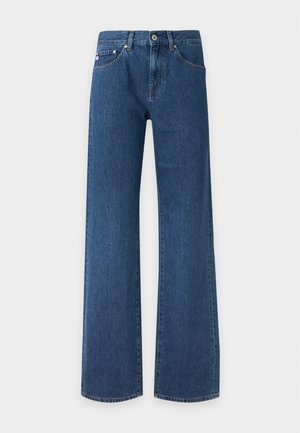 Femme aux cheveux foncés portant un t-shirt marron, un jean bleu, un pull crème noué à la taille, des lunettes de soleil foncées, des chaussures noires, et tenant un sac à main crème.