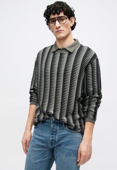 Πλεκτό πουλόβερ με μαύρο και γκρι ζιγκ-ζαγκ μοτίβο, με μυτερό γιακά, μακριά μανίκια και πλεκτές μανσέτες. Συνδυάζεται με μπλε τζιν.