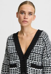 Melna un balta adīta kardigāna jakas ar V-veida kakla izgriezumu, ar strukturētu rakstu, ribu apdari un divām priekšējām kabatām. Priekšpusē trīs pogas.