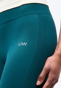 Τυρκουάζ αθλητικό κολάν χωρίς ραφές με υφασμένες πλευρικές λεπτομέρειες και λογότυπο στο μηρό, εμφανίζεται στο ισχίο και το χέρι ενός ατόμου.