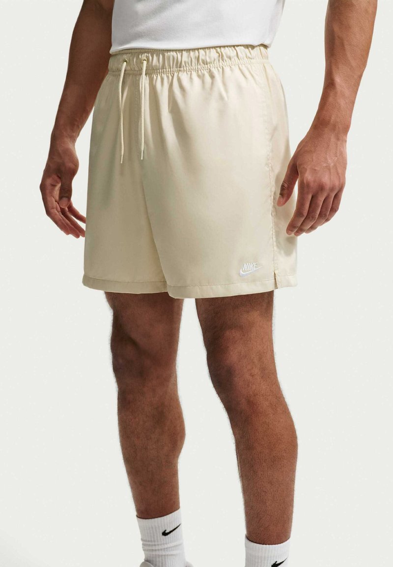 Man draagt beige Nike short met elastische tailleband en trekkoord, witte Nike sokken, staand tegen een effen achtergrond.
