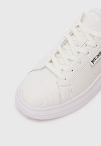 Sneaker bianca con loghi texture sulla punta, lacci bianchi e etichetta "Just Cavalli" sul lato.