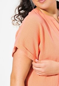 Top rosa pesca a maniche corte realizzato in tessuto testurizzato, caratterizzato da una vestibilità rilassata e sottili dettagli stropicciati, che evidenziano la leggerezza del tessuto.
