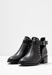 Botas de tobillo de cuero negro con tachuelas plateadas, correas con hebillas y tacones cuadrados sobre una superficie blanca con un fondo liso.