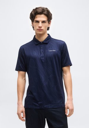 Νέος άνδρας που φοράει μπλε ναυτικό πόλο της Calvin Klein με διακριτικά κυματιστά σχέδια, στέκεται μπροστά σε απλό ανοιχτό φόντο.