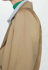 Cappotto trench beige con una superficie liscia, caratterizzato da un colletto prominente e tasche nascoste. Indossato sopra un dolcevita verde e una camicia a righe.