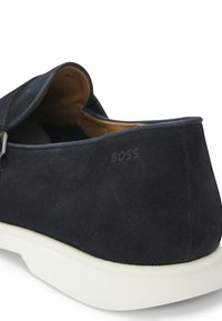 Dark blue suede shoe with a smooth texture, featuring a white rubber sole. Subtle embossed logo on the heel and an open back design.