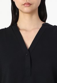 Top negro con escote en forma de V y un detalle de un solo botón. Tejido suave y texturizado con un ajuste relajado. Diseño minimalista, sin patrones ni acentos.