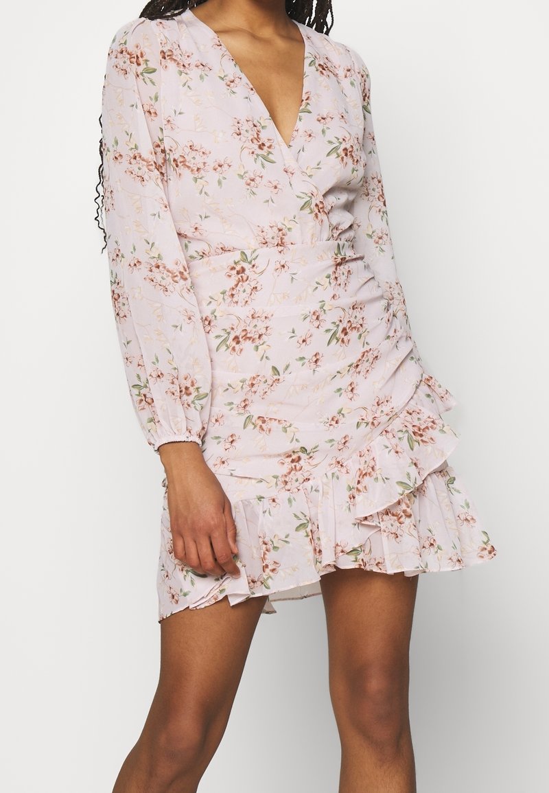 Femme portant une robe portefeuille fleurie rose clair à manches longues et ourlet à volants, debout devant un fond uni.