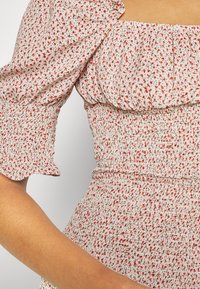 Haut à manches bouffantes fleuri en tissu léger ; caractérisé par un col volanté, une taille smockée et un motif de petites fleurs rouges et noires sur un fond beige.