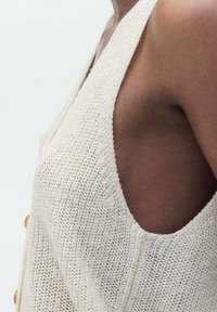 Zblízka osoba v bezrukávovém, pleteném svetru krémové barvy s velkými otvory na paže a viditelnými knoflíky po straně.