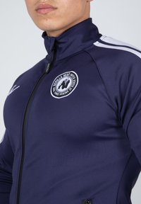 Marine sportjack met een ritssluiting, hoge kraag, zijkanten in kleur en een ronde witte logo met een beer. Glad, rekbaar polyester materiaal.