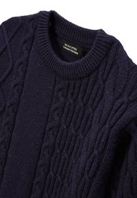 Pull en maille torsadée bleu foncé avec un col rond et un motif texturé avec des points en relief. Étiquette visible à l'intérieur du col.