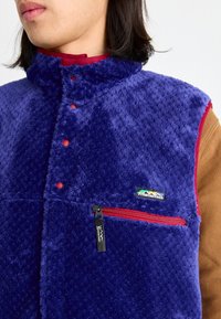 Fleece vest in purple with a zip pocket and contrasting red accents. Features a high collar and button closure. Brand logo displayed.