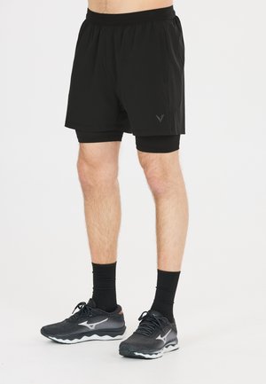 Onderlichaam van een persoon die zwarte gelaagde hardloopshorts, zwarte sokken en zwarte sportschoenen draagt op een effen witte achtergrond.