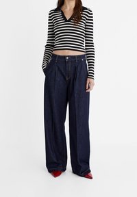Czarnobiały w pasy prążkowany crop top z dekoltem w kształcie litery V, połączony z ciemnoniebieskimi szerokimi spodniami dżinsowymi i czerwonymi szpilkami w szpic.