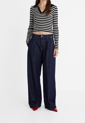 Czarnobiały w pasy prążkowany crop top z dekoltem w kształcie litery V, połączony z ciemnoniebieskimi szerokimi spodniami dżinsowymi i czerwonymi szpilkami w szpic.