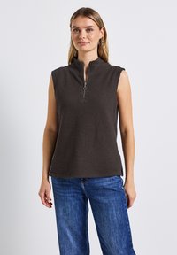 Mörkbrun ärmlös topp med hög krage och dragkedja fram, med en texturerad, våffel-liknande mönster och sidofickor. Parat med blå jeans.
