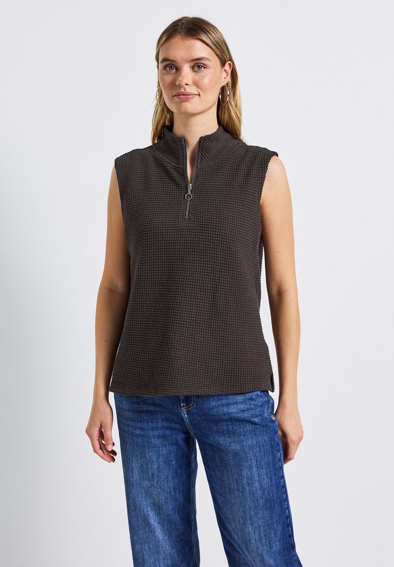 Mörkbrun ärmlös topp med hög krage och dragkedja fram, med en texturerad, våffel-liknande mönster och sidofickor. Parat med blå jeans.