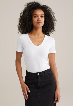 Woman with curly hair wearing a white V-neck T-shirt tucked into a high-waisted black denim skirt, standing with hand on hip.