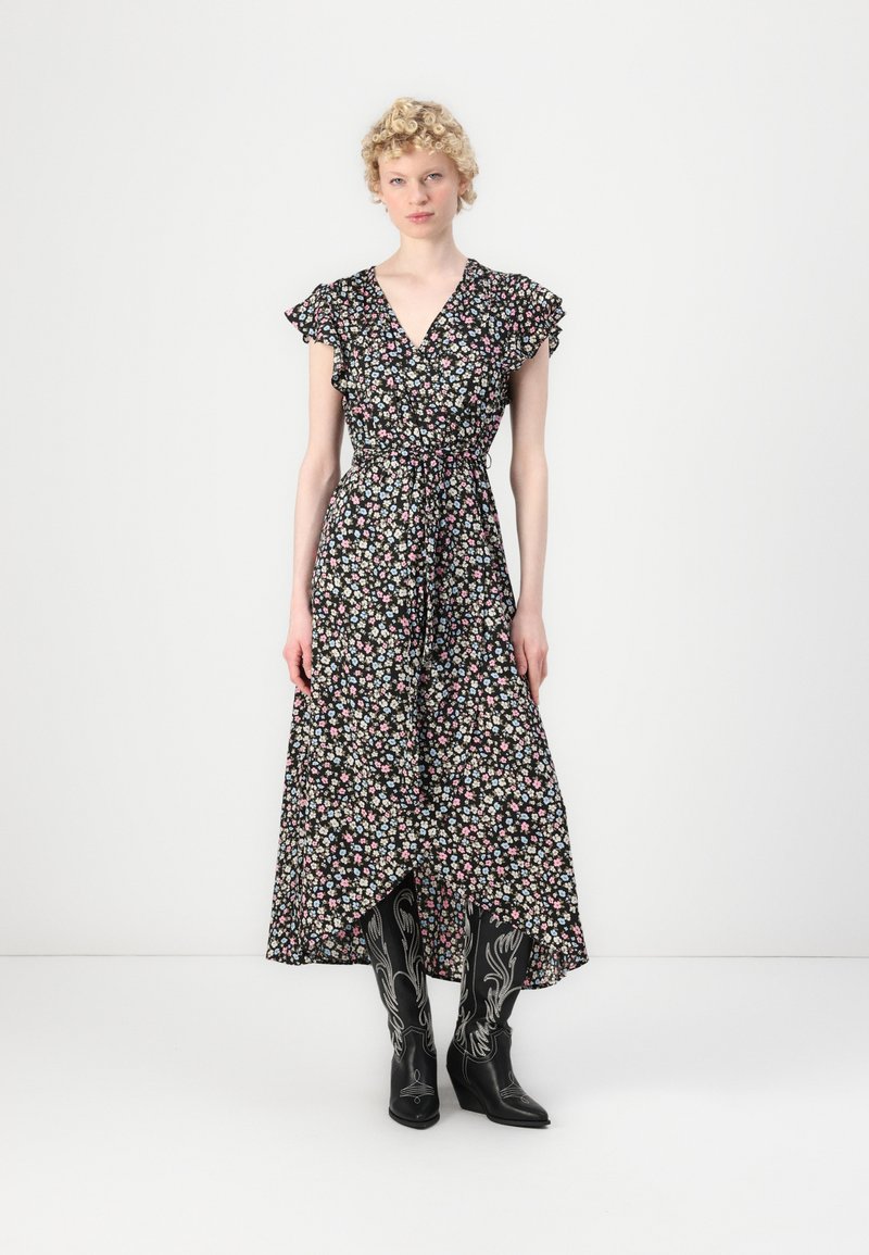Robe portefeuille à fleurs avec de courtes manches volantées, fond noir, fleurs roses et bleues, taille ceinturée et ourlet longueur cheville. Associée à des bottes de cowboy noires.