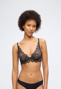 Γυναίκα με σγουρά μαλλιά που φοράει μαύρο δαντελένιο σουτιέν και ταιριαστά μαύρα εσώρουχα, στέκεται μπροστά σε απλό φωτεινό φόντο.