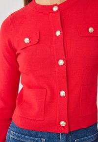 Roter Cardigan mit gerippter Textur, Rundhalsausschnitt, vier vorderen Taschen und goldfarbenen Knöpfen. Fitted Design, sichtbare Nähte und glattes Material.