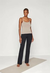 Naine seisab paljajalu beežil joogatamatil, seljas beež särgike ja mustad jooga püksid, lihtsa valge taustaga.